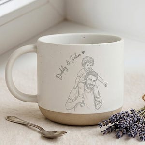 May include: A white ceramic mug with a speckled finish and a tan base, featuring a grey line-art illustration of a person carrying a child on their shoulders. The text "Daddy & John" with a small heart icon is printed in grey script on the side.