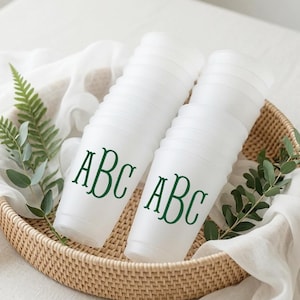 May include: A stack of translucent white plastic cups featuring the dark green monogram letters ABC printed on the front. The cups are arranged in two columns within a round woven basket lined with white fabric and decorated with green leaves.