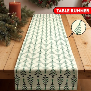 May include: Cream-colored table runner with a repeating pattern of green Christmas trees. The trees have a black star at the base. The words "TABLE RUNNER" are in a red banner. The text "FUCK TRUMP" is on the trees.