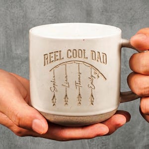 May include: Cream-colored ceramic mug with the text "REEL COOL DAD" and the names Sophia, Liam, Thomas, and Emily. The mug features a fishing theme with fish illustrations and a handle.