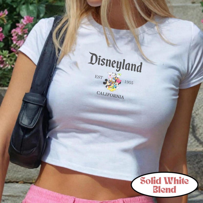 Op de afbeelding: Witte crop top met het Disneyland-logo in zwart schrift, met Mickey Mouse en vrienden. Het shirt bevat de tekst "EST 1955 CALIFORNIA". Het shirt is een effen witte mix.