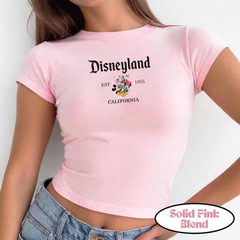 Op de afbeelding: Een lichtroze t-shirt met het woord "Disneyland" in een decoratief lettertype. Daaronder staat de tekst "EST 1955 CALIFORNIA" met een afbeelding van Disney-personages. Het shirt is een effen roze mix.