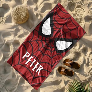 Puede incluir: Una toalla de playa roja con un dise&ntilde;o de telara&ntilde;a negra y formas de ojos blancos, con el nombre "PETER" en letras blancas. La toalla est&aacute; sobre arena, con un sombrero de paja y sandalias marrones.