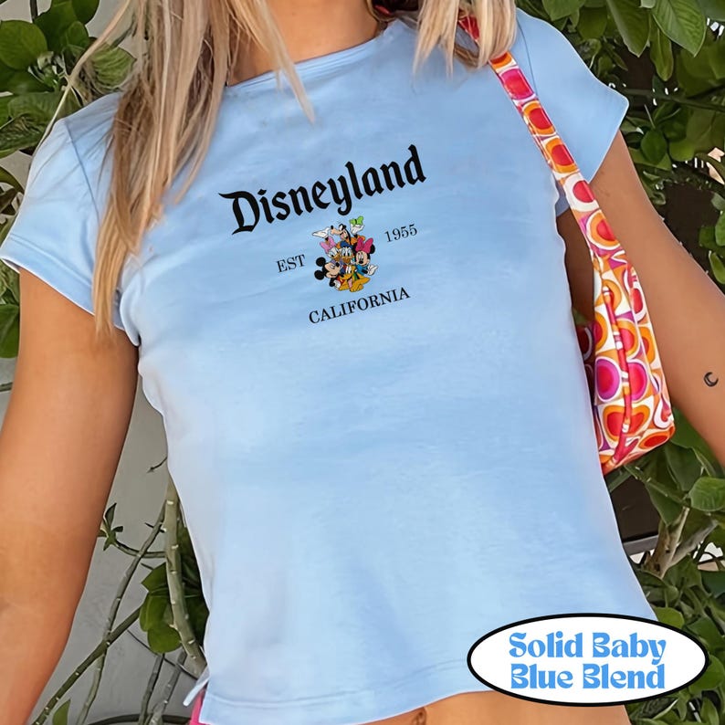 Op de afbeelding: Lichtblauw T-shirt met het woord "Disneyland" in zwart schrift, de tekst "EST 1955 CALIFORNIA" en een afbeelding van Mickey en Minnie Mouse. Het shirt is een effen babyblauwe mix.