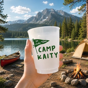 May include: A translucent plastic cup featuring a green graphic of a pennant flag with the text '26 Lake Tahoe' and the words 'CAMP KAITY' printed in a bold, green, sans-serif font on the front surface of the cup.