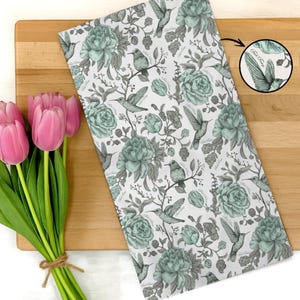 May include: A white kitchen towel with a floral and hummingbird pattern in shades of gray and teal. The towel is lying on a wooden cutting board next to a bouquet of pink tulips.
