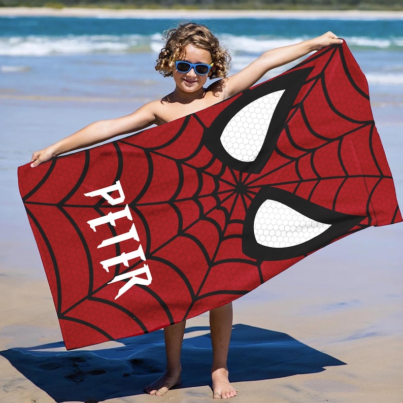 Puede incluir: Toalla de playa roja con un dise&ntilde;o de telara&ntilde;a negra y grandes ojos blancos, que recuerdan a una m&aacute;scara de superh&eacute;roe. El nombre "PETER" est&aacute; impreso en blanco. La toalla se sostiene en una playa.