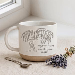 May include: A cream-colored ceramic mug with a dark gray illustration of a willow tree featuring a heart carved with the letters A+H. The text reads I "WILLOW" WAYS Love You and the date 08.11.2017. The base of the mug is unglazed tan clay.