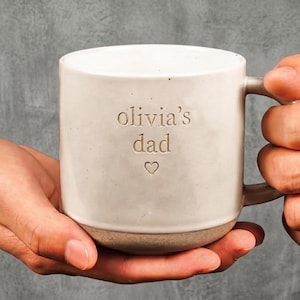May include: A cream-coloured ceramic mug with a speckled finish and a handle on the right side. The front of the mug features the engraved text "olivia's dad" in lowercase letters, positioned above a small, simple engraved heart symbol.