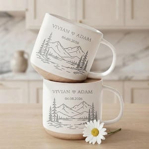 May include: Two off-white ceramic mugs stacked vertically, each featuring a dark grey line art illustration of mountains, pine trees, and a lake. The text VIVIAN HEART ADAM and the date 06.08.2026 are printed above the landscape design.