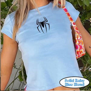 May include: Light blue t-shirt with a black spider graphic. The shirt has short sleeves and a crew neck. The text "SOLID BABY BLUE BLEND" is in a white oval at the bottom of the image.