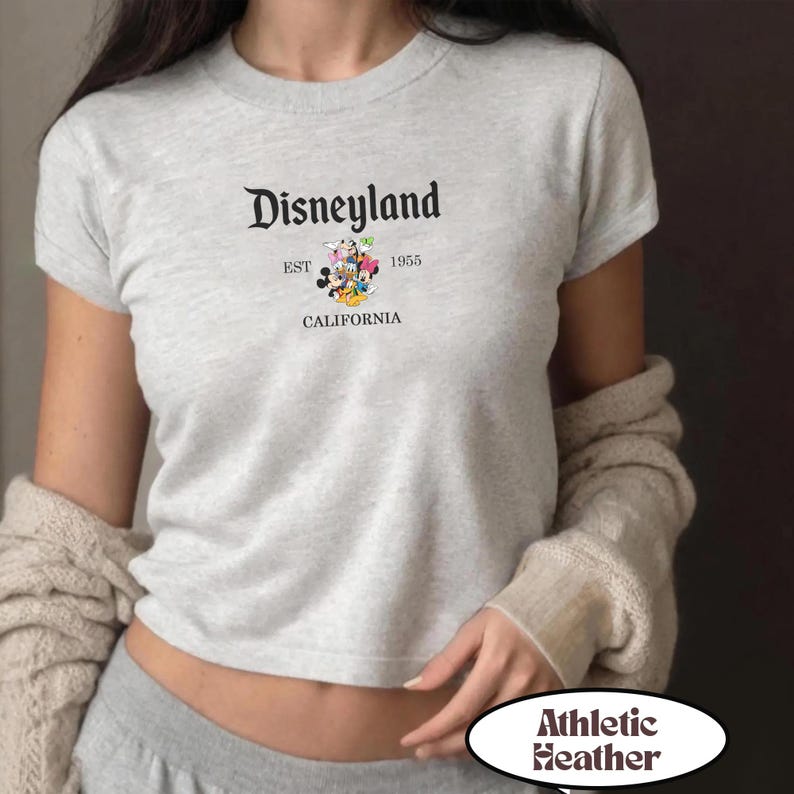 Op de afbeelding: Lichtgrijs Athletic Heather T-shirt met het Disneyland-logo. Het logo bevat de tekst "Disneyland" boven "EST 1955 CALIFORNIA" en een afbeelding van Mickey en Minnie Mouse, Donald Duck en Goofy.