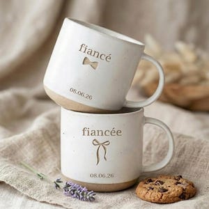 May include: Two off-white ceramic mugs with speckled textures. The top mug features the word fiancé, a small bow tie icon, and the date 08.06.26. The bottom mug features the word fiancée, a ribbon bow icon, and the date 08.06.26.