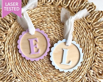 May include: Two wooden tags with scalloped edges, one is light blue with a white letter L and the other is light purple with a white letter E. The tags are on a woven straw surface.