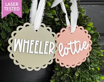 May include: Two scalloped-edge ornaments with white ribbon hangers. One is sage green with the name "WHEELER" in white block letters. The other is peach with the name "lottie" in white script. The text "DESIGNS BY DANA DIGITAL" is at the top.