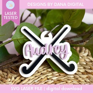 Puede incluir: Adorno blanco cortado con láser con palos de hockey negros cruzados y el nombre "Audrey" en escritura rosa. También se ve el texto "DESIGNS BY DANA DIGITAL" y "SVG LASER FILE | digital download".
