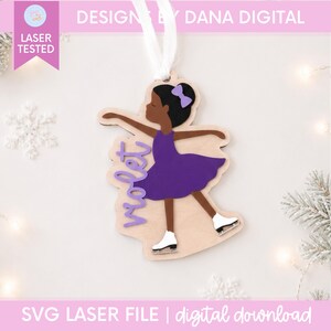 May include: A wooden ornament featuring a dark-skinned figure ice skating in a purple dress with a matching purple hair bow. The name Violet is written in purple script on the left side. A white ribbon is attached to the top for hanging.