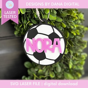 Op de afbeelding: Een ronde voetbal sleutelhanger met de naam "NORA" in roze letters. De sleutelhanger is zwart en wit met een zilverkleurige metalen lus. De woorden "DESIGNS BY DANA DIGITAL" en "SVG LASER FILE | digital download" zijn ook zichtbaar.