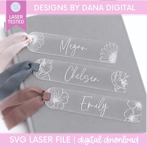 May include: Three rectangular clear acrylic bookmarks with rounded ends, each engraved with a floral design and a name: Megan, Chelsea, and Emily. Each bookmark features a frayed fabric ribbon tied through a hole at one end in light pink, blue, and mauve.
