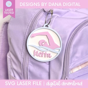 May include: A circular light blue and white acrylic bag tag featuring a pink silhouette of a swimmer in a pool. The name Kierra is written in pink script lettering below the swimmer. The tag is attached to a light purple backpack with a metal loop.