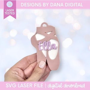 May include: A dusty rose ballet slipper cutout featuring a cutout design and the name Ella in light purple script lettering. The object includes a small circular hole at the top for hanging and is labelled as a laser tested SVG laser file.