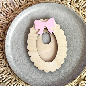 May include: A wooden oval-shaped frame with a scalloped edge and a pink bow on top. The frame has a cutout in the center in the shape of an oval.