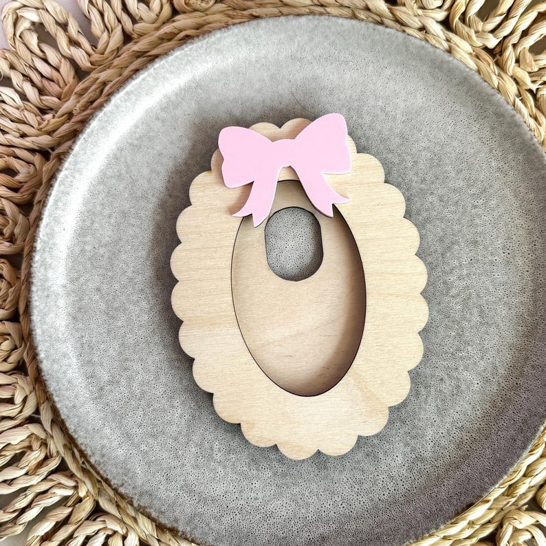 May include: A wooden oval-shaped frame with a scalloped edge and a pink bow on top. The frame has a cutout in the center in the shape of an oval.