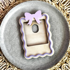May include: A wooden picture frame with a scalloped edge and a purple bow on top. The frame is made of light wood and has a cutout in the center for a photo.