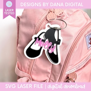 Pode incluir: Uma etiqueta de bolsa em acrílico preto e branco em forma de par de sapatos com o nome Molly escrito em letras cursivas cor-de-rosa. A etiqueta está presa a uma mochila rosa claro com um cabo metálico prateado e tem um contorno branco.