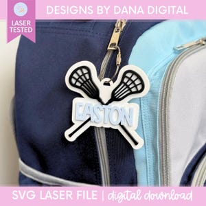 May include: A white and black lacrosse stick bag tag featuring the name EASTON in light blue block letters. The tag is attached to a navy blue and light blue backpack with a silver metal cable loop. The design includes two crossed lacrosse sticks.