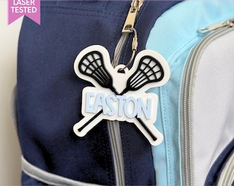 May include: A white and black lacrosse stick bag tag featuring the name EASTON in light blue block letters. The tag is attached to a navy blue and light blue backpack with a silver metal cable loop. The design includes two crossed lacrosse sticks.