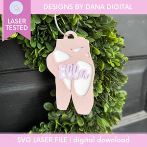 May include: A pink laser-cut ballet shoe ornament with the name "Ella" in lavender. The ornament is hanging in front of a green wreath. The text "DESIGNS BY DANA DIGITAL" and "LASER TESTED" are visible.