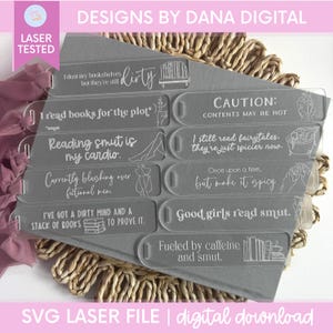 May include: A collection of clear acrylic bookmarks with humorous quotes in white lettering. The bookmarks have a rectangular shape with a curved top and a small hole. The phrases include "Reading smut is my cardio" and "Good girls read smut."