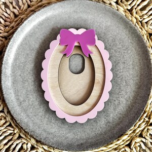 May include: A wooden oval-shaped frame with a pink scalloped edge and a pink bow on top. The frame is designed to hold a photo or other small item.