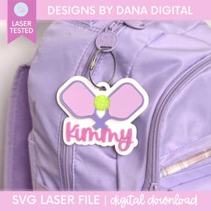 May include: A custom acrylic bag tag featuring a purple pickleball paddle graphic and a yellow pickleball, with the name Kimmy written in pink script lettering. The tag is attached to a light purple backpack with a silver metal wire loop.