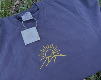 Può includere: Una maglietta blu scuro con un design ricamato giallo di montagna e sole. La maglietta ha un girocollo e una piccola etichetta con le parole "Thank You". La maglietta è appoggiata sull'erba verde.