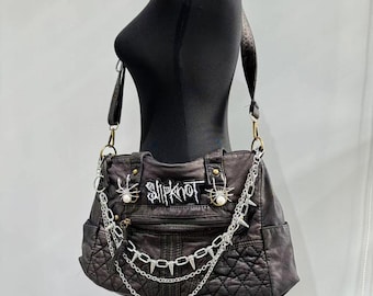 Könnte beinhalten: Schwarze Lederschultertasche mit gestepptem Design und silbernen Ketten. Die Tasche hat einen großen gestickten Aufnäher mit der Aufschrift "Slipknot" und zwei silberne Spinnenanhänger.