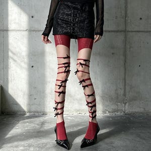 May include: Red tights featuring a sheer design with a darker red garter-style top section and multiple thin red ribbons wrapped around the legs, each accented with small black bows. The tights are paired with black pointed-toe high heels.