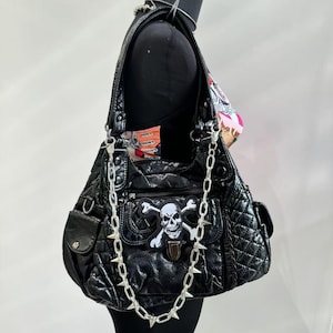 Puede incluir: Bolso de hombro negro acolchado con un parche de calavera y huesos cruzados. El bolso tiene una cadena plateada con púas y múltiples bolsillos. El bolso se muestra en un maniquí negro.