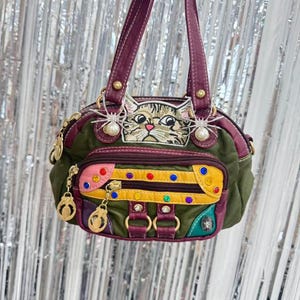 Puede incluir: Un bolso único con un diseño de cara de gato. El bolso presenta un cuerpo verde con detalles burdeos, un bolsillo con cremallera rosa y amarillo, y abalorios decorativos. La cara del gato está bordada y hay adornos en forma de araña a cada lado.