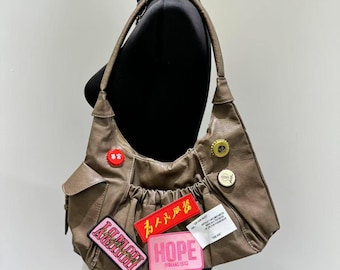 May include: A brown leather shoulder bag with multiple patches and pins. The bag has a slouchy design with a front pocket and a long strap. Patches include the words "HOPE" and "GLORY".