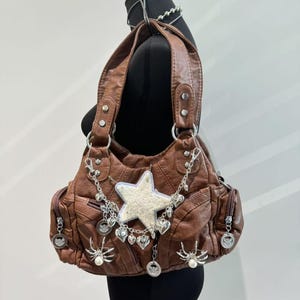 Può includere: Una borsa a mano in pelle marrone con dettagli argentati, tra cui un ciondolo a forma di stella, ciondoli a forma di cuore e ciondoli a forma di ragno.
