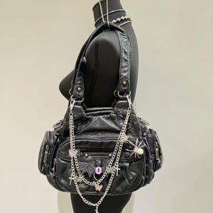 May include: A black leather handbag with multiple pockets and zipper closures. The bag is adorned with a silver chain and charms, including a bat, crosses, butterflies, and a spider.