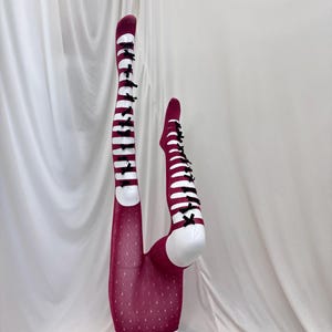 Peut inclure: Une paire de collants magenta foncé présentant une rangée verticale de découpes horizontales sur le devant de chaque jambe, agrémentée de petits nœuds en ruban noir au centre de chaque ouverture. La partie inférieure a un motif losange.