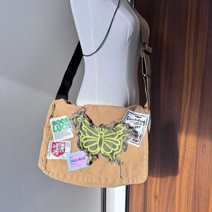 May include: A brown canvas messenger bag with a green butterfly patch and various other patches, including "Good Luck" and "Coca Cola Blue".