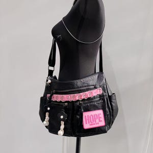 Puede incluir: Bolso de hombro de cuero negro con detalles en rosa y un parche rosa que dice "HOPE" en letras blancas. El bolso tiene varios bolsillos y un adorno de flor decorativo.