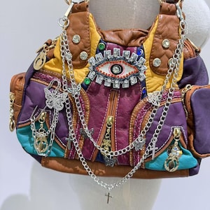 Puede incluir: Un bolso de cuero multicolor con un gran dise&ntilde;o de ojo en la parte delantera. El bolso tiene varias cadenas y abalorios, incluyendo mariposas, cruces y una peque&ntilde;a llave.