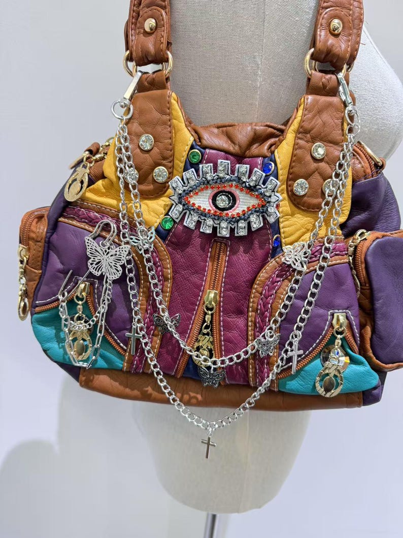 Puede incluir: Un bolso de cuero multicolor con un gran dise&ntilde;o de ojo en la parte delantera. El bolso tiene varias cadenas y abalorios, incluyendo mariposas, cruces y una peque&ntilde;a llave.