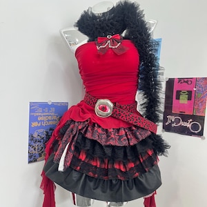 Puede incluir: Un vestido rojo sin tirantes con un detalle de tul negro en el cuello, un lazo rojo con cadena plateada y un cinturón de estampado de leopardo con hebilla plateada en forma de ojo. La falda tiene volantes superpuestos de encaje y tela.