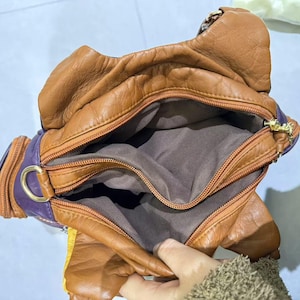 Puede incluir: Un bolso de mano de cuero marr&oacute;n con forro morado y varios compartimentos. El bolso tiene cierre de cremallera y una hebilla dorada.